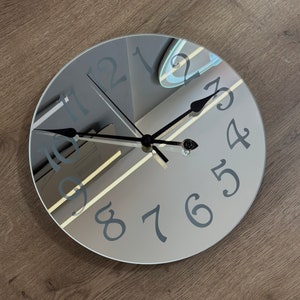 Op de afbeelding: Een ronde, gespiegelde klok met zwarte wijzers en zilveren cijfers. De wijzerplaat van de klok is reflecterend en toont een reflectie van de kamer.