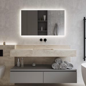 Può includere: Un mobile da bagno moderno con un piano in bianco e un mobiletto grigio. Il mobile da bagno ha un lavabo integrato e uno specchio rettangolare grande con una luce intorno al bordo. Ci sono due bottiglie di lozione e un barattolo di crema sul piano, e una pila di asciugamani sul mobiletto.