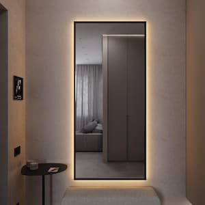 May include: A rectangular mirror with a black frame and warm LED lighting. The mirror is mounted on a light-colored wall above a gray upholstered bench. A small black side table and a framed picture are also visible.