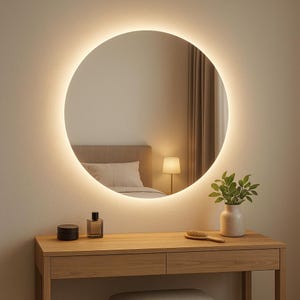 Può includere: Uno specchio rotondo illuminato è appeso sopra un mobile in legno. Lo specchio ha una luce calda e brillante attorno al bordo. Sul mobile ci sono un barattolo nero, una bottiglia di profumo, una spazzola e un vaso con foglie verdi. La stanza ha una palette di colori neutri.