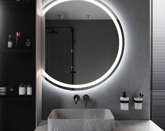 May include: A modern bathroom scene featuring a large, round mirror with a bright, illuminated border. Below the mirror is a rectangular, gray stone sink with black fixtures. The walls are gray, and the overall aesthetic is minimalist and contemporary.