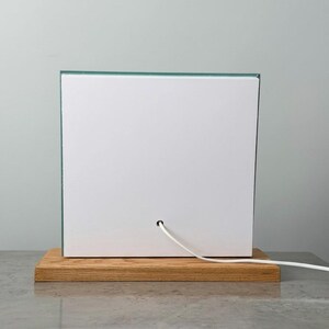 Op de afbeelding: Een moderne, vierkante lamp met een witte voorkant en een heldere glazen rand. De lamp staat op een rechthoekige houten basis. Een wit netsnoer komt uit de achterkant.