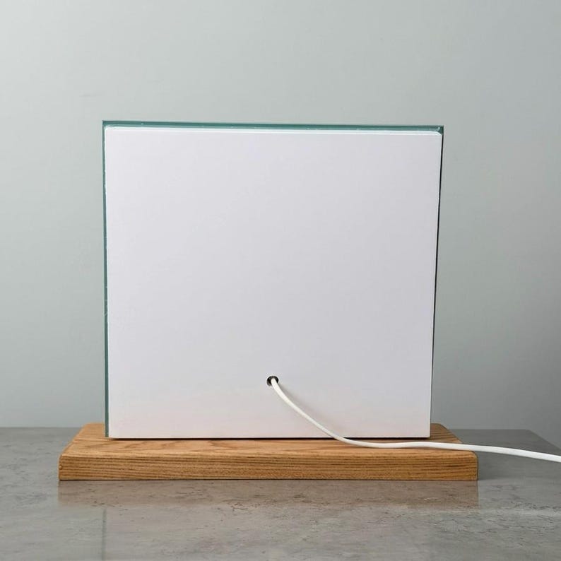 Op de afbeelding: Een moderne, vierkante lamp met een witte voorkant en een heldere glazen rand. De lamp staat op een rechthoekige houten basis. Een wit netsnoer komt uit de achterkant.