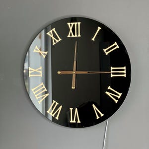 Puede incluir: Un reloj de espejo negro con números romanos y manecillas doradas.