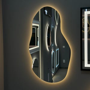 May include: A modern, irregularly shaped mirror with a white LED light around the perimeter. The mirror reflects other mirrors with white LED lights, creating a geometric pattern.