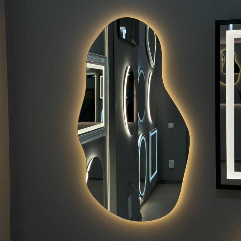 May include: A modern, irregularly shaped mirror with a white LED light around the perimeter. The mirror reflects other mirrors with white LED lights, creating a geometric pattern.
