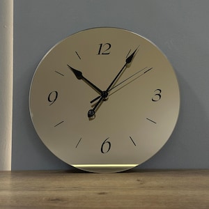 Op de afbeelding: Een ronde, goudkleurige wandklok met zwarte cijfers en wijzers. De klok heeft een minimalistisch ontwerp met een subtiele, verlichte strook aan de onderkant. De klok is bevestigd aan een grijze muur.