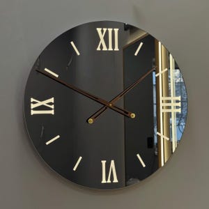 May include: A round, black wall clock with Roman numerals and wooden hands. The clock face is reflective, and the numerals are in a light gold color. The clock hands are dark brown with gold accents.