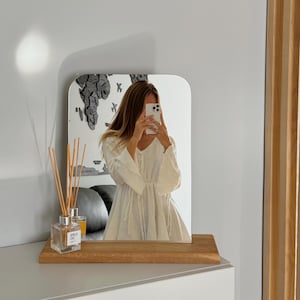 May include: A rectangular mirror with rounded corners sits on a wooden shelf. The mirror reflects a person holding a phone. A glass diffuser with reeds and a small glass of liquid are on the shelf.