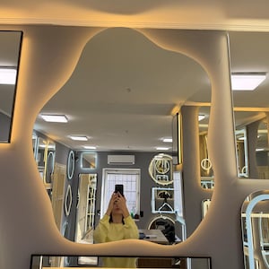 May include: A large, irregularly shaped mirror with a soft, organic outline. The mirror is surrounded by a warm, glowing light. The image shows a person holding a phone up to their face, reflecting the room and other mirrors.