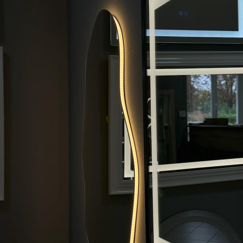 May include: A modern, wavy-shaped mirror with a thin, warm white LED light strip around the perimeter. The mirror is mounted on a black wall and reflects a room with a window.