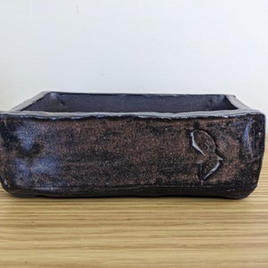 May include: A rectangular ceramic planter with a dark, textured finish in shades of black and deep brown. The vessel features thick, uneven walls with a rustic, hand-formed appearance and a small, subtle leaf-shaped indentation on the front side.