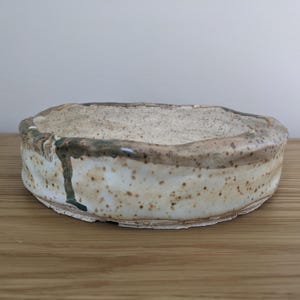 Op de afbeelding: Een ondiepe, ronde keramische kom met een onregelmatige, met de hand gevormde rand. De buiten- en binnenkant zijn voorzien van een gespikkeld gebroken wit glazuur met bruine en donkergroene druppelaccenten langs de textuurrijke, ongelijke rand.