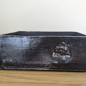 Puede incluir: Una maceta rectangular de cerámica con un esmalte marrón oscuro, brillante y texturizado. La superficie exterior presenta un sutil detalle en relieve con forma de concha en la parte frontal. El recipiente tiene paredes gruesas.