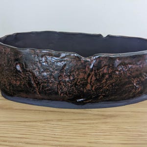 May include: An oval ceramic bowl with a dark, textured exterior featuring a mottled black and copper-brown glaze. The rim is uneven and organic in shape, while the interior is finished in a smooth, solid dark purple or deep black glaze.