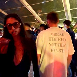Puede incluir: Una camiseta blanca con el texto "BREAK HER BED, NOT HER HEART" impreso en la espalda. La camiseta la lleva una persona entre la multitud, con una mujer con gafas de sol en primer plano.