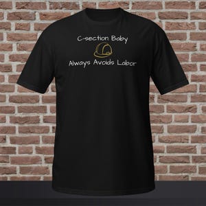 Puede incluir: Camiseta negra con el texto blanco "C-section Baby Always Avoids Labor" y un gráfico de casco de construcción amarillo. La camiseta se muestra sobre un fondo de pared de ladrillo.