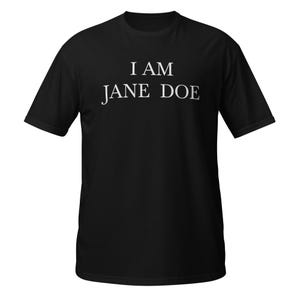 May include: A black short-sleeved t-shirt featuring the white text "I AM JANE DOE" printed in a serif font across the chest. The garment has a crew neckline and a standard fit, presenting a plain black background with centered white lettering.