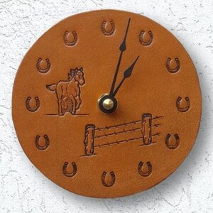 May include: A brown leather clock with a horse and fence design. The clock face is surrounded by horseshoe outlines.