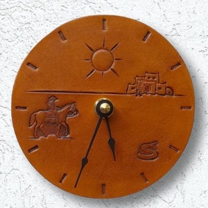 May include: A brown leather clock with a sun, a building, a rider on a horse, and a snake design. The clock hands are black and the clock face is white.