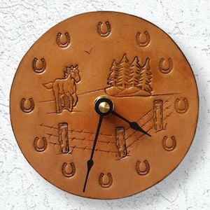 May include: A round, brown leather clock with a Western theme. The clock face features a horse, a fence, trees, and horseshoes. The clock hands are black, and the clock is mounted on a white textured wall.