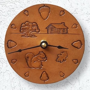May include: A brown leather clock with a squirrel, a cabin, a tree, and acorns carved into the face. The clock hands are black.