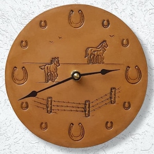 May include: A brown leather clock with a horse and horseshoe design. The clock face has a gold-colored hour and minute hand. The clock is surrounded by a brown leather frame.