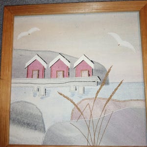 以下が含まれることがあります： 海岸の風景を描いた額入りのアートワーク。白い屋根の3つのピンク色のコテージが灰色の構造の上にあります。2羽の白いカモメが空を飛んでいます。アートワークは、明るい茶色の木製フレームに入っています。