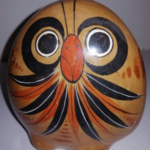 May include: A hand-painted ceramic owl figurine. The owl has large black and white eyes, an orange beak, and a brown body with orange and black feather details. The owl has a rounded shape and three small feet.