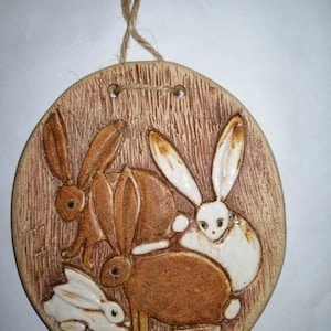 Könnte beinhalten: Ein Keramik-Wandbehang mit brauner und weißer Glasur, der drei Kaninchen zeigt. Die Kaninchen sind in einer Gruppe angeordnet, mit einem weißen Kaninchen in der Mitte und zwei braunen Kaninchen auf beiden Seiten. Der Wandbehang ist oval geformt und hat einen braunen Hintergrund mit einer Holzmaserung. Der Wandbehang ist unten rechts mit "Marita" signiert.