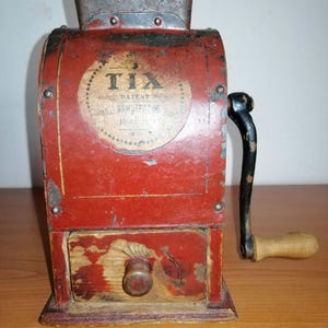 Könnte beinhalten: Eine antike, handbetriebene Mühle aus rotem Metall mit einer Holzschublade und einem Griff. Auf der Vorderseite der Mühle steht das Wort "TIX", zusammen mit dem Wort "PATENT" und anderem Text. Die Mühle wird wahrscheinlich zum Mahlen von Kaffeebohnen oder Gewürzen verwendet.