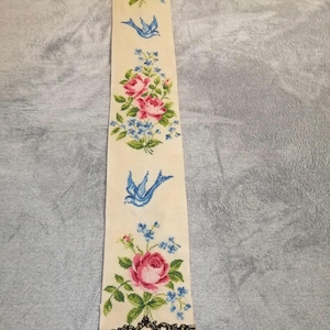 Könnte beinhalten: Cremefarbener, bestickter Wandbehang mit rosa Rosen, blauen Vergissmeinnicht und blauen Vögeln. Dekorative Metallverzierungen säumen die Ober- und Unterkante. Das Blumen- und Vogelmuster wiederholt sich über die gesamte Länge.