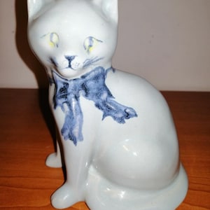 May include: A ceramic figurine of a light blue cat wearing a blue bow. The cat is sitting with its tail curled around its legs.