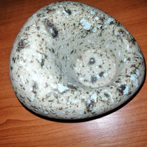 Könnte beinhalten: Eine gesprenkelte, ovale Keramikschale mit einem abgerundeten, offenen Innenraum. Die Schale hat eine hellbeige Grundfarbe mit dunkelbraunen und hellblauen Sprenkeln. Der Innenraum ist glatt und die Außenseite hat ein strukturiertes Aussehen.