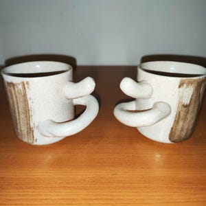 Può includere: Due tazze in ceramica dal design unico. Le tazze bianco sporco presentano dettagli a pennellate marroni e manici curvi distintivi. Sono posizionate fianco a fianco su una superficie di legno, evidenziando la loro qualità artigianale e il loro stile artistico.