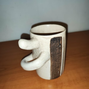 Peut inclure: Une tasse en céramique faite à la main avec une conception de poignée unique. La tasse est blanc cassé avec des rayures verticales marron et un accent rectangulaire. La poignée a une forme incurvée distinctive. La tasse est adaptée au café, au thé ou à d'autres boissons.