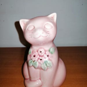 May include: A pink ceramic cat figurine with a floral design around its neck. The cat is sitting with its paws tucked under its body and has a closed-eye expression.