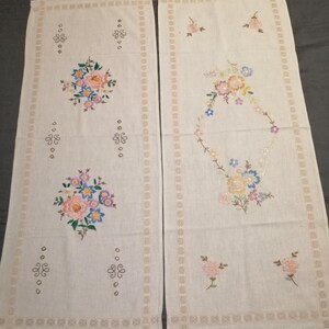 Könnte beinhalten: Zwei weiße Tischläufer aus Leinen mit gestickten Blumendesigns. Die Designs zeigen rosa, blaue, gelbe und grüne Blumen.