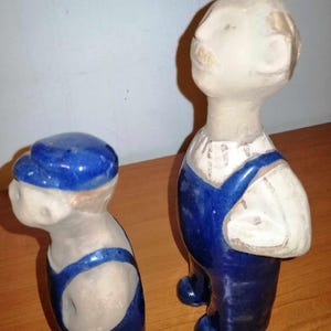 Può includere: Due figurine in ceramica di persone che indossano tute blu. La figurina a sinistra indossa un berretto blu. Entrambe le figurine hanno volti bianchi e sono dipinte in stile rustico.