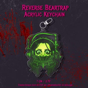 May include: Acrylic keychain with a green and black illustration of a character from the horror film series, Saw. The keychain is shaped like a reverse beartrap, with the text "REVERSE BEARTRAP ACRYLIC KEYCHAIN". The keychain is 7 cm in size.