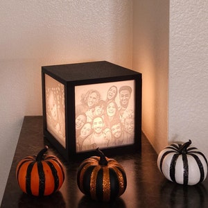 May include: A black cube-shaped lamp with a photo of a group of people inside. The lamp is lit and casting a warm glow. Three decorative pumpkins are on a dark surface in front of the lamp.