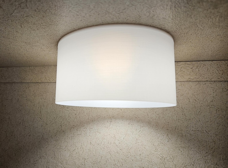 Może przedstawiać: Biała, cylindryczna lampa sufitowa. Światło ma miękki, rozproszony blask i jest zamontowane na teksturowanym, neutralnym kolorystycznie suficie. Design oprawy jest prosty i nowoczesny, odpowiedni dla r&oacute;żnych styl&oacute;w wnętrz. Średnica wynosi około 30 cm.