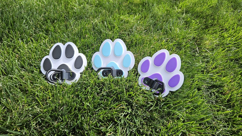May include: Three paw print shaped keychains with black, blue, and purple paw prints on a white background. Each keychain has a black metal clip.