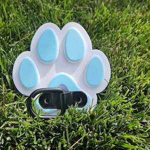 May include: A white paw print shaped dog leash clip with a blue paw print design. The clip is made of black metal.