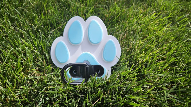 May include: A white paw print shaped dog leash clip with a blue paw print design. The clip is made of black metal.