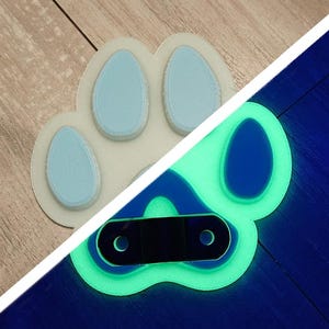 May include: A white paw print with blue toes glows green under a black light. The paw print has a black metal ring attached to it.