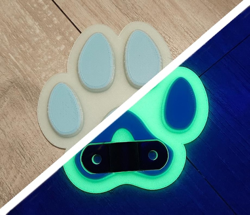 May include: A white paw print with blue toes glows green under a black light. The paw print has a black metal ring attached to it.