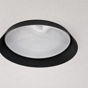 May include: A white, oval-shaped ceiling light fixture with a black trim. The light has a small on/off switch and is set into a white ceiling. The fixture is designed for interior lighting.