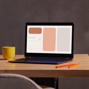 Puede incluir: Un portátil abierto que muestra un calendario de noviembre de 2026 con una paleta de colores neutros. Una taza amarilla y dos bolígrafos naranjas están sobre un escritorio de madera. El portátil muestra un diseño de calendario digital.