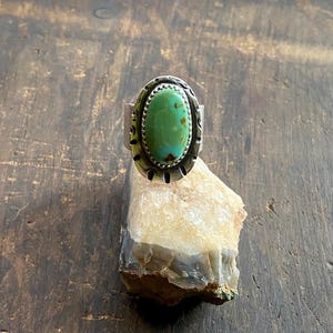 May include: A silver ring with an oval, light green turquoise stone. The stone has brown inclusions and is set in a decorative bezel. The ring is displayed on a light-colored rock against a dark wood surface. The ring's design has a vintage or Southwestern aesthetic.